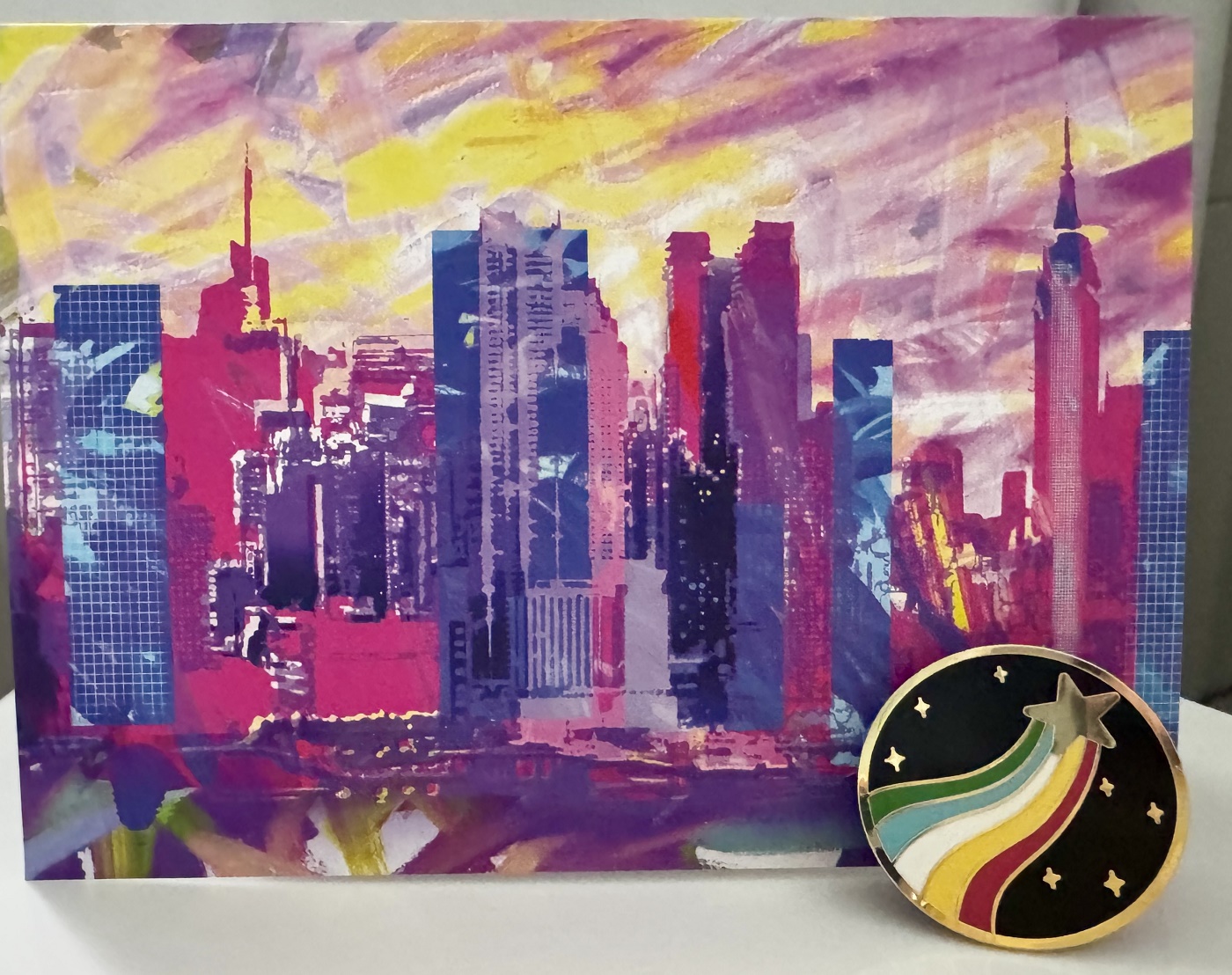 A vibrant visual painting of New York City’s skyline, created by Annie who was born with cerebral palsy. In front of it is a version of Annie’s progressive Disability Pride flag which is the shooting star.